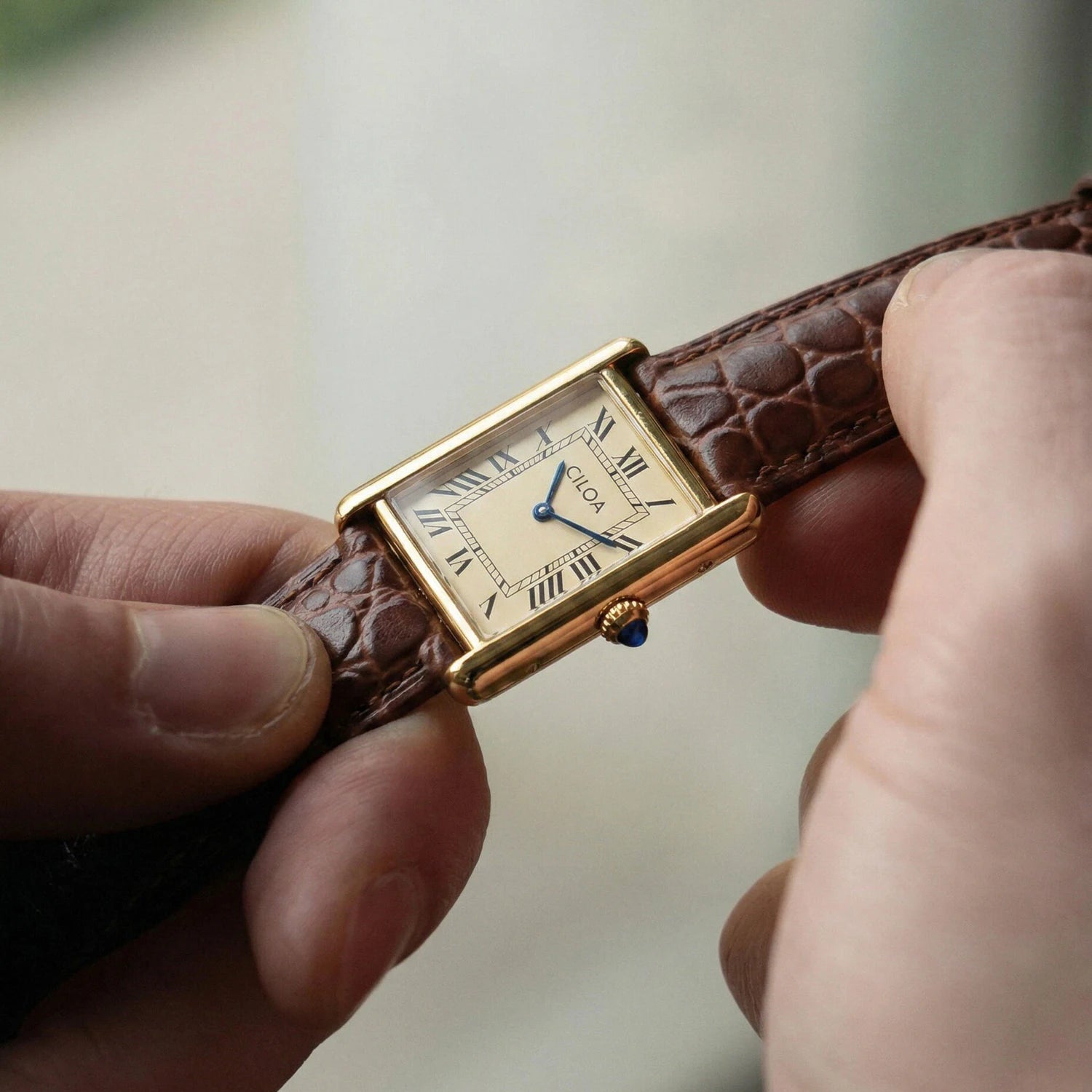 Angled close-up of the gold CILOA square wristwatch, highlighting the blue gem cabochon on the crown and the brown leather strap texture