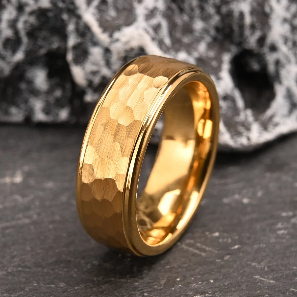 Close-up display of the gold classic hammered tungsten carbide ring standing upright on a rustic wet stone slab background