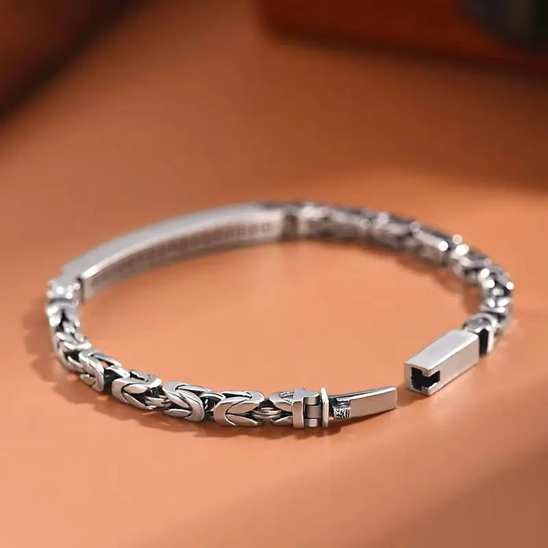 Open silver men's ID bracelet showing the secure clasp mechanism and intricate woven Byzantine chain on a warm brown surface.