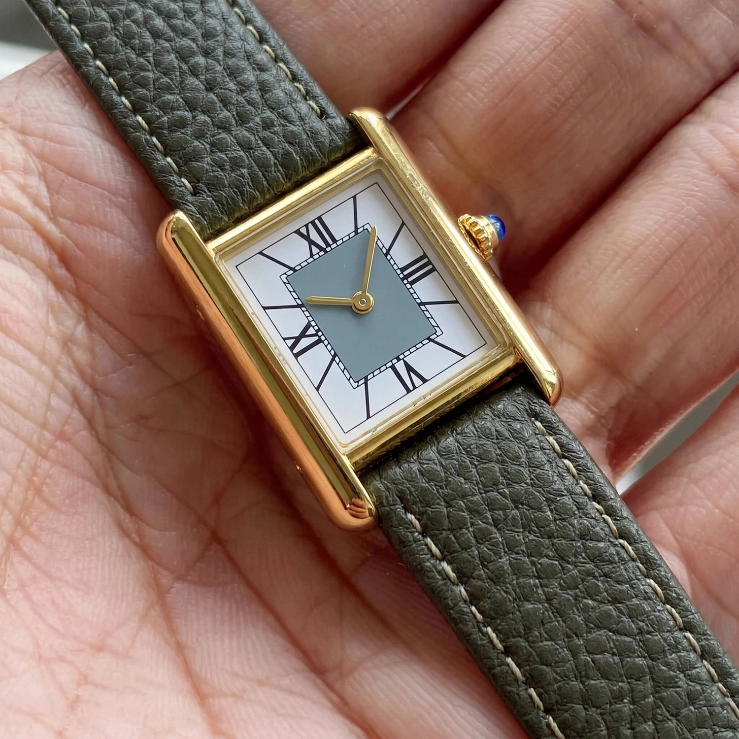 Elegant rectangular gold watch with Roman numerals and a green leather strap resting in the palm of a hand to showcase its size and elegant profile.