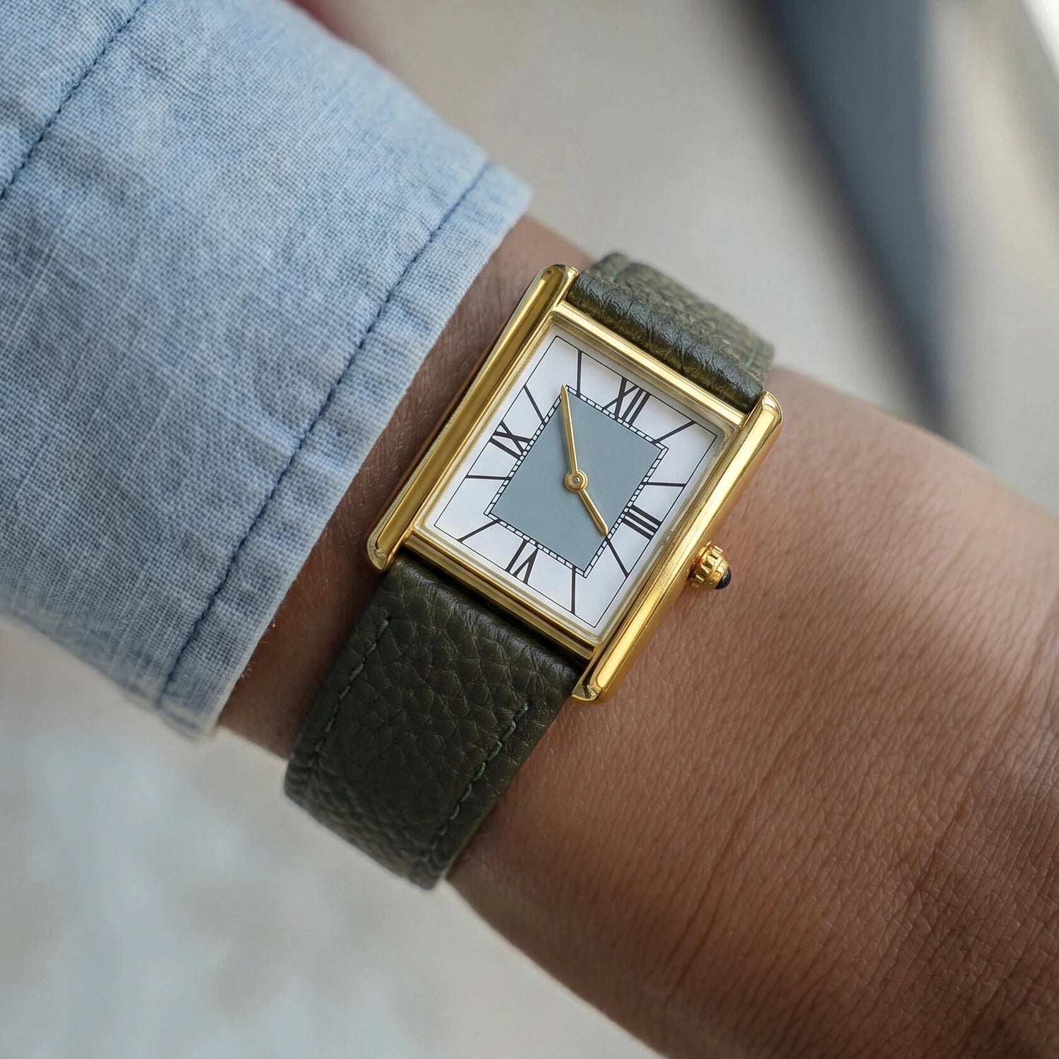 Lifestyle shot of a classic rectangular gold watch with a grey dial and green leather strap worn on the wrist with a blue sleeve.