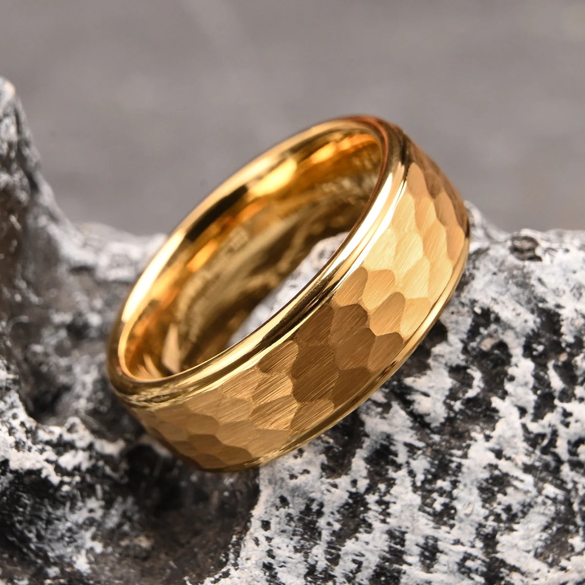A luxury gold-plated hammered tungsten ring balanced on a textured grey volcanic rock, highlighting its heavy-duty scratch resistance
