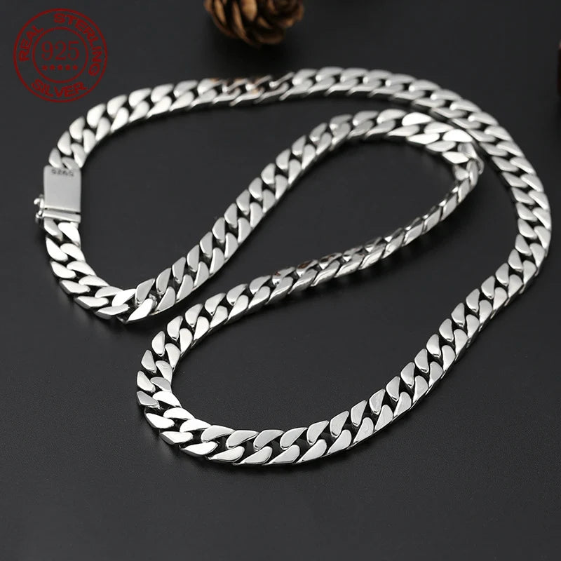 Heavy silver flat curb chain necklace curled naturally on a dark grey surface.