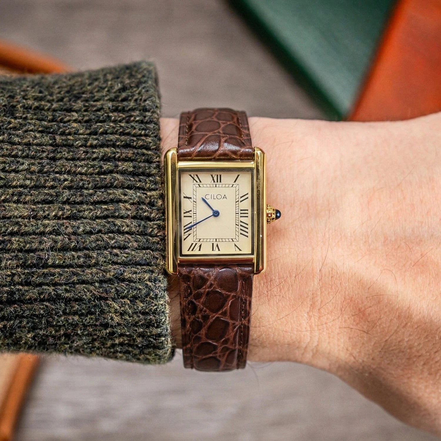 A man wearing an olive green ribbed knit sweater showcasing the luxury gold CILOA retro square watch with its brown alligator leather strap