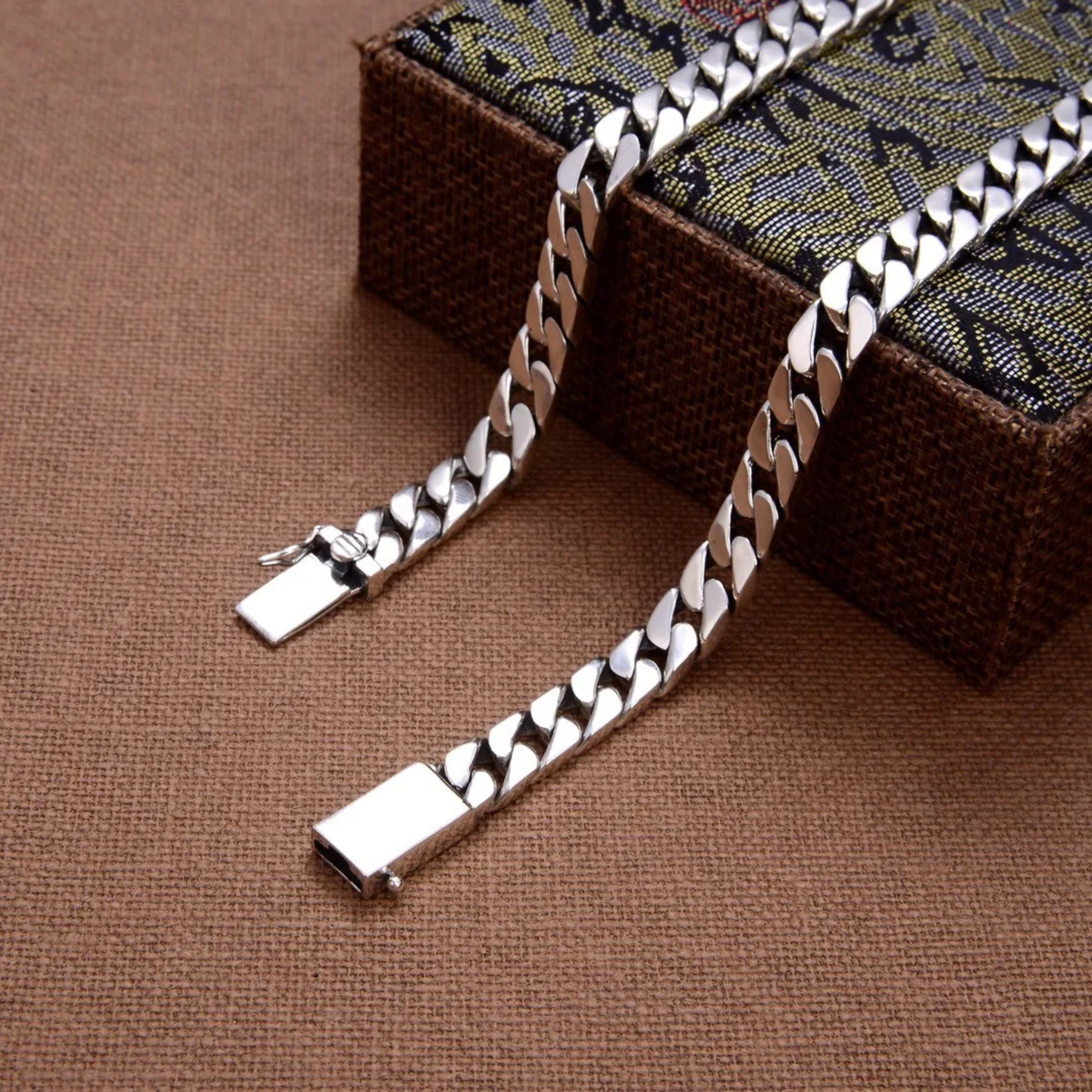 Silver curb chain necklace with an open box clasp resting next to a patterned box on a warm brown surface.