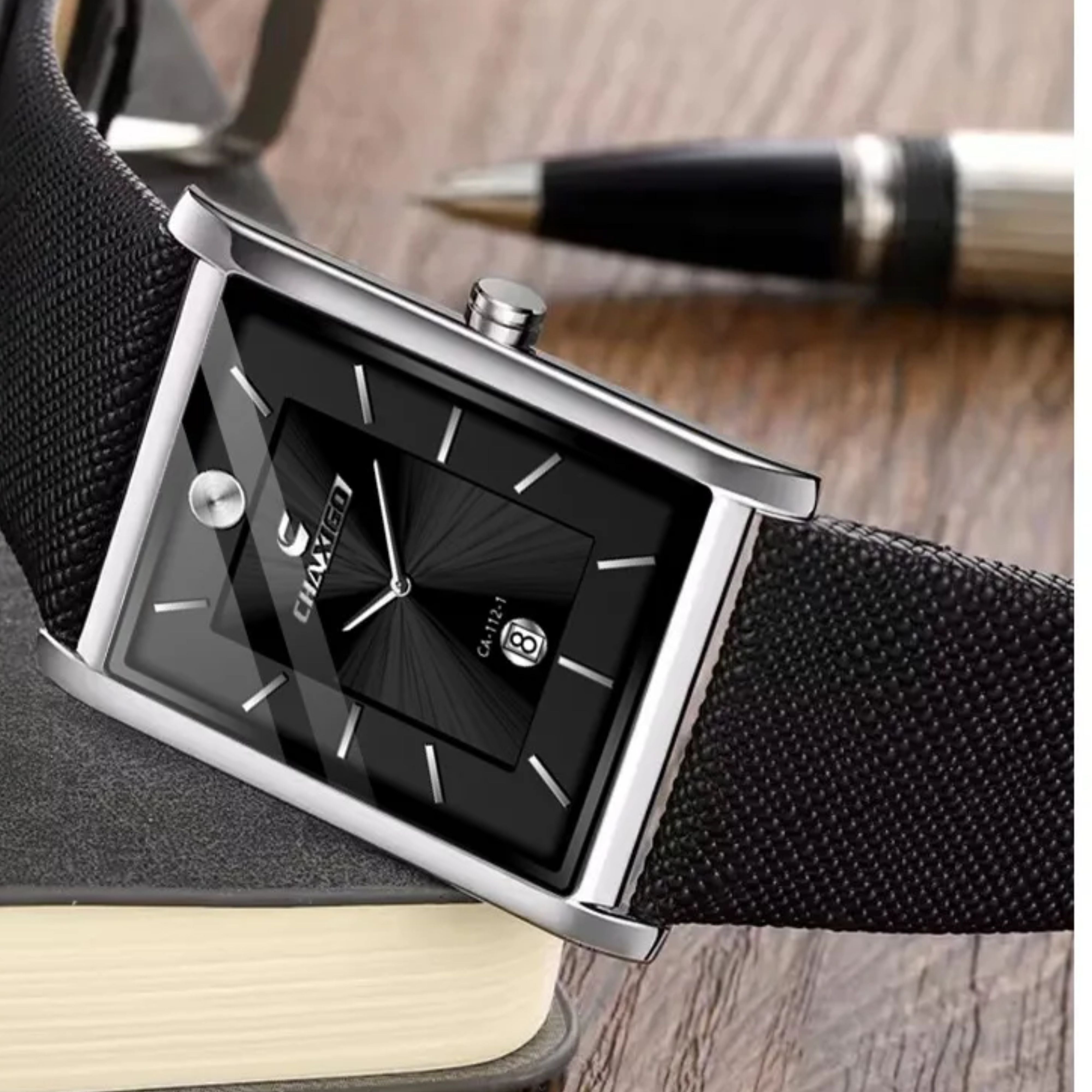 Silver tank style men's watch with a black dial resting on a wooden desk next to a business pen and notebook.