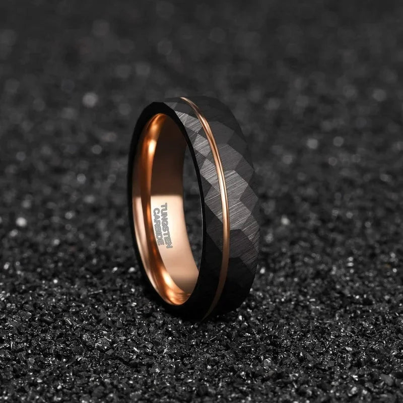 Studio product presentation of the Tigrade black and rose gold hammered tungsten ring standing upright on a textured dark asphalt surface